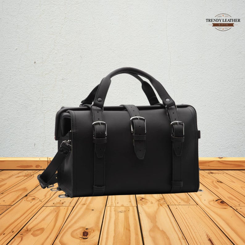 May include: Black leather satchel bag with a top handle and adjustable shoulder strap. The bag features three buckle straps on the front and a flap closure. The bag is sitting on a wooden surface.