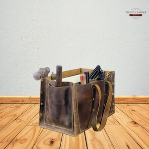 Può includere: Una borsa porta attrezzi in pelle marrone con più tasche e un manico. La borsa contiene vari attrezzi, tra cui un martello, uno scalpello e delle pinze. La borsa è realizzata in pelle invecchiata e ha un aspetto vintage. La borsa è appoggiata su un pavimento in legno.