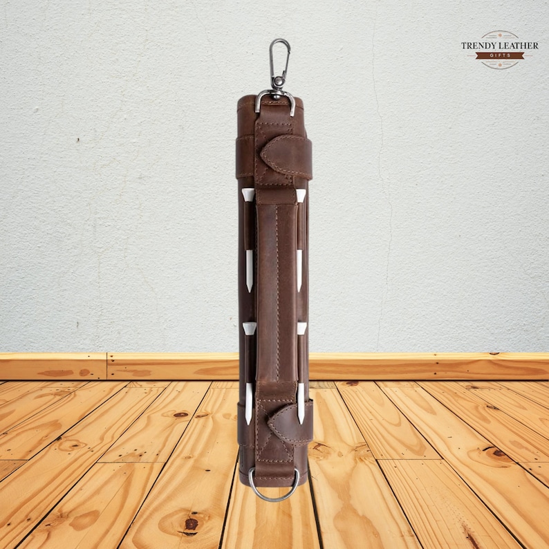 May include: A brown leather golf club carrier with white accents and a silver-colored clasp. The carrier has a handle and a ring at the bottom. The Trendy Leather logo is in the top right corner.