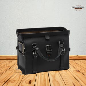 May include: A black leather satchel with a top handle and multiple silver buckles. The bag has a side pocket and a flap closure. The Trendy Leather logo is visible in the top right corner. The bag is sitting on a wooden surface.