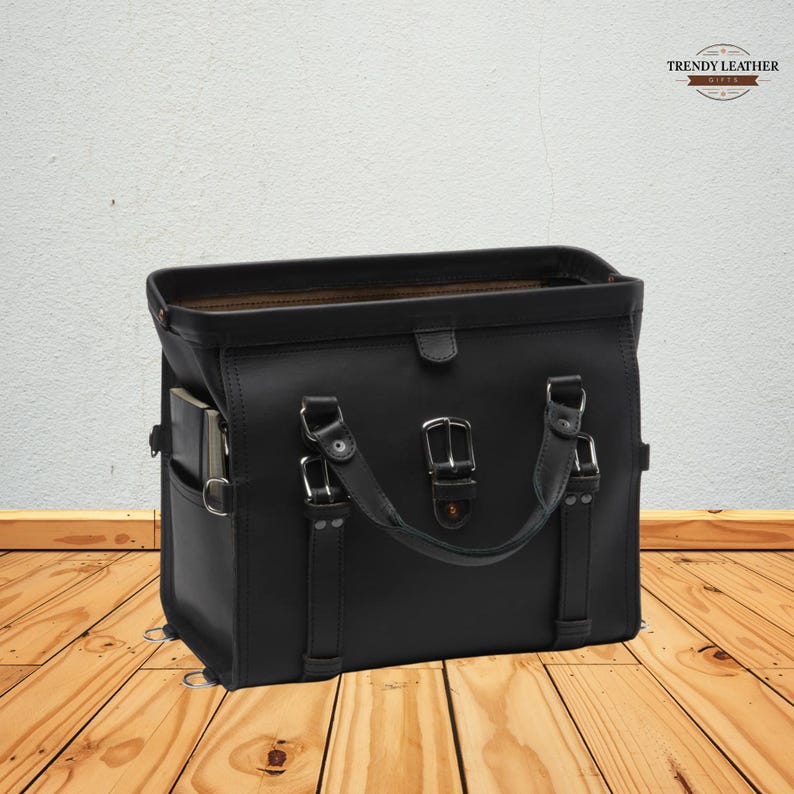 May include: A black leather satchel with a top handle and multiple silver buckles. The bag has a side pocket and a flap closure. The Trendy Leather logo is visible in the top right corner. The bag is sitting on a wooden surface.