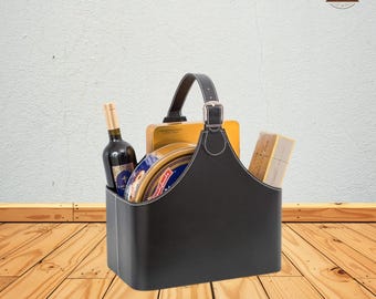 May include: A black leather gift basket with a handle, white stitching, and a silver buckle. The basket contains a bottle of wine, a tin of cookies, and a box of crackers. The basket is on a wooden surface.