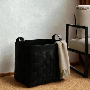 May include: A black woven storage basket with a solid top rim and two integrated handles. A beige knit throw blanket is draped over the right side of the basket. The basket features a square-patterned weave design and a smooth, matte finish.