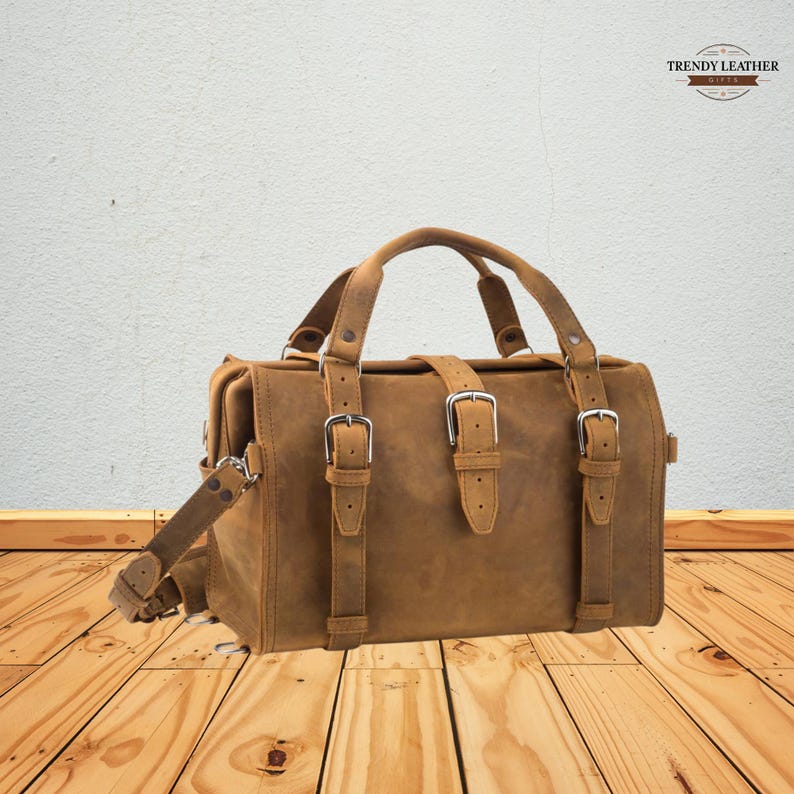 May include: A brown leather duffel bag with silver buckles and straps. The bag has a top handle and a shoulder strap. The bag is made of distressed leather and has a vintage look. The bag is sitting on a wooden surface.