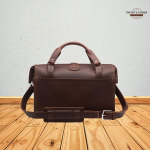 May include: A brown leather briefcase with a top handle and a detachable shoulder strap. The bag has a rectangular shape with a front pocket and a buckle closure. The bag has the words "TRENDY LEATHER" in the top right corner.