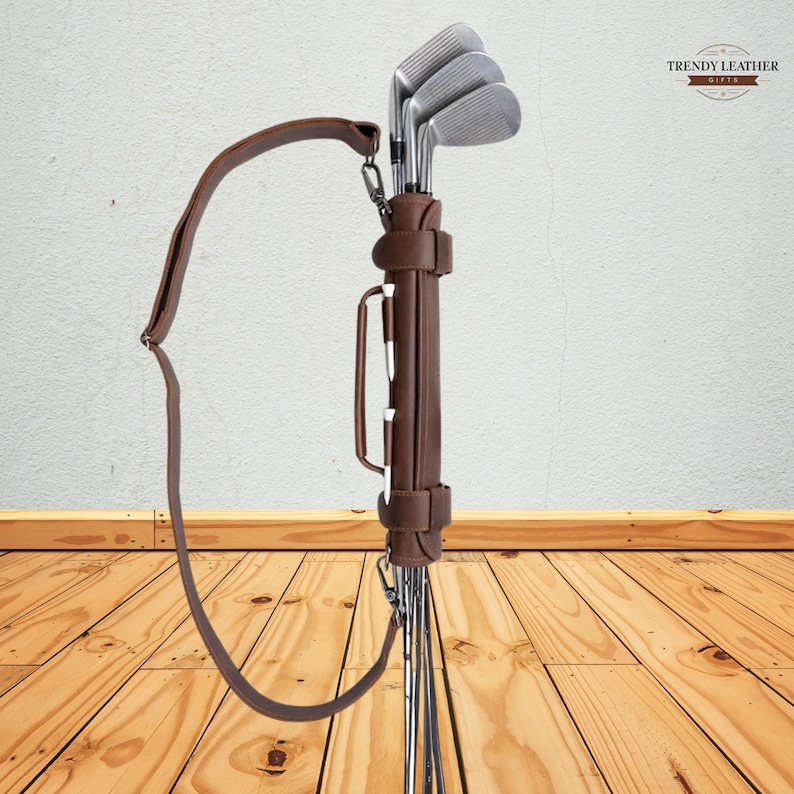 May include: A brown leather golf club carrier with a shoulder strap. The carrier holds several golf clubs with silver heads. The carrier has a handle and is branded with the text "TRENDY LEATHER GIFTS".
