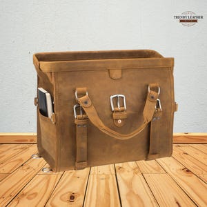 May include: A brown leather satchel bag with a rectangular shape. The bag features a top handle, side pocket, and adjustable straps with silver buckles. A notebook is visible in the side pocket. The bag has the text "TRENDY LEATHER" on the top right.