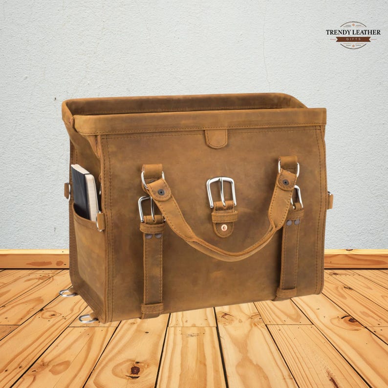 May include: A brown leather satchel bag with a rectangular shape. The bag features a top handle, side pocket, and adjustable straps with silver buckles. A notebook is visible in the side pocket. The bag has the text "TRENDY LEATHER" on the top right.