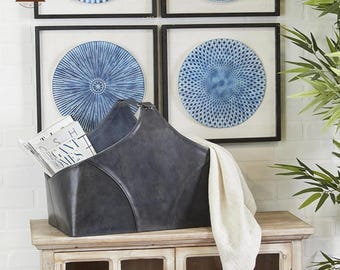 May include: A dark gray leather storage basket with a handle, holding magazines and a cream-colored cloth. The basket sits on a wooden cabinet, with framed blue and white decorative plates on the wall behind it. The Trendy Leather logo is visible.