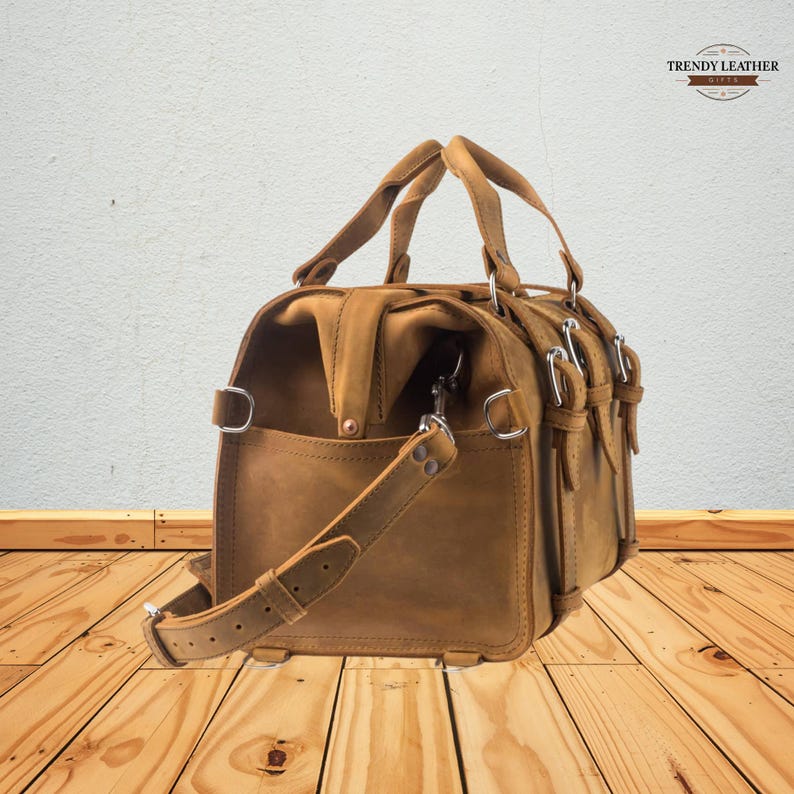 May include: A brown leather satchel bag with a top handle and a detachable shoulder strap. The bag has multiple compartments, metal buckles, and a front pocket. The bag is made of distressed leather and has a vintage look.