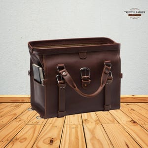 May include: A brown leather briefcase with a top handle and silver buckles. The bag has a side pocket and a notebook inside. The Trendy Leather logo is in the top right corner. The bag is sitting on a wooden surface.