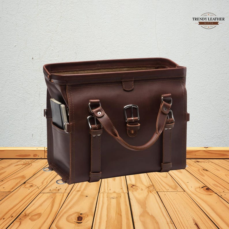 May include: A brown leather briefcase with a top handle and silver buckles. The bag has a side pocket and a notebook inside. The Trendy Leather logo is in the top right corner. The bag is sitting on a wooden surface.