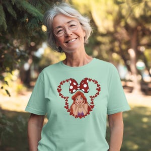 A smiling woman wearing a green t-shirt with a cartoon character and a bow on it, standing in a park.