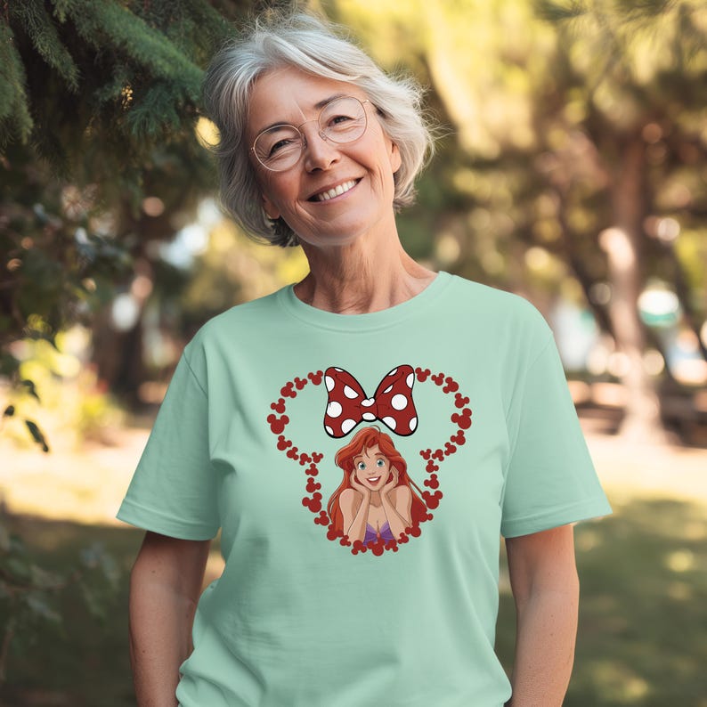 A smiling woman wearing a green t-shirt with a cartoon character and a bow on it, standing in a park.
