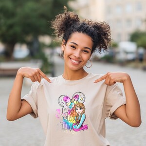 A smiling young woman wearing a t-shirt with a cartoon character on it, pointing at the shirt while standing outdoors.