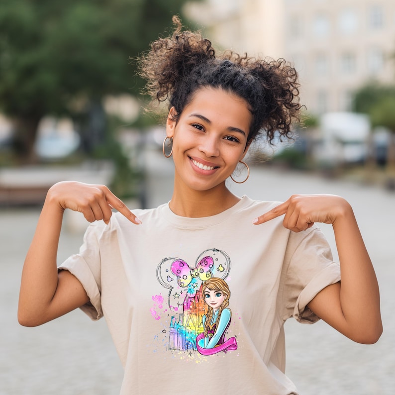 A smiling young woman wearing a t-shirt with a cartoon character on it, pointing at the shirt while standing outdoors.