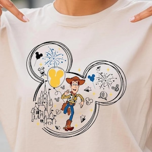 A smiling young woman wearing a t-shirt with a Mickey Mouse design and pointing at it.