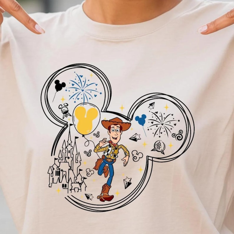 A smiling young woman wearing a t-shirt with a Mickey Mouse design and pointing at it.