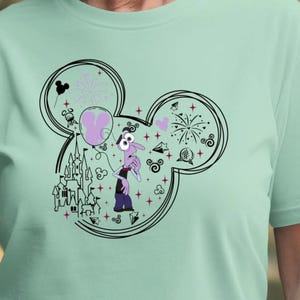 May include: A light green t-shirt featuring a black Mickey Mouse head outline with a cartoon character inside. The character is purple and has a worried expression. The design includes a castle, fireworks, and a balloon.
