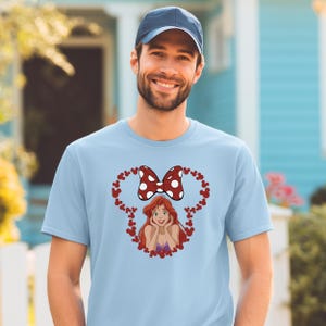 A smiling man wearing a blue baseball cap and a light blue t-shirt with a cartoon character and heart design.