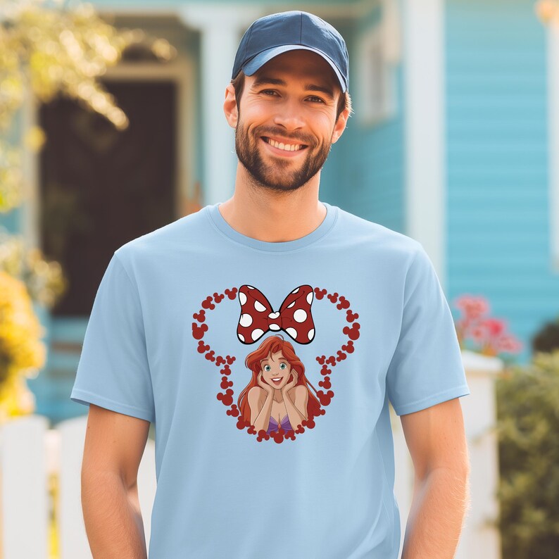 A smiling man wearing a blue baseball cap and a light blue t-shirt with a cartoon character and heart design.