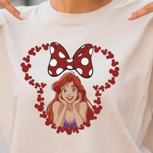 A smiling young woman wearing a t-shirt with a cartoon character on it, pointing at the shirt while standing outdoors.