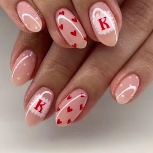 Puede incluir: Uñas ovaladas de color rosa claro con varios diseños. Algunas uñas presentan pequeños corazones rojos, otras tienen un borde blanco con corazones rojos, y algunas tienen un borde blanco con la letra "K" en rojo. Unas pocas uñas tienen lunares blancos.