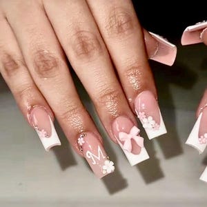May include: Artificial nails with a square shape. The nails have a pink base with white tips, decorated with small white flowers, a pink bow, and the letter "M". Some nails are embellished with tiny, sparkling details.