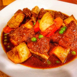 Op de afbeelding: Een close-up van een stevige stoofpot geserveerd in een witte kom. De stoofpot bevat stukken vlees, goudbruine aardappelen, rode wortelen en groene erwten, allemaal ondergedompeld in een rijke, roodbruine saus. Het gerecht wordt gepresenteerd op een wit bord.