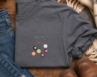 May include: A gray t-shirt with an embroidered pool-themed design. The design includes crossed pool cues, a white cue ball, and several numbered billiard balls. The text "RACK 'EM UP" is above the design. The shirt is displayed with blue jeans and brown boots.