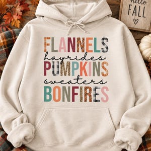May include: A cream-colored hooded sweatshirt featuring a front graphic with the words FLANNELS, hayrides, PUMPKINS, sweaters, and BONFIRES. The letters are styled in various autumn-themed patterns including plaid, leopard print, and solid muted colors.