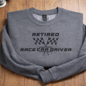 May include: A gray sweatshirt with the text "RETIRED RACE CAR DRIVER" in black. The design includes a checkered flag graphic. The sweatshirt is folded on a wooden surface.