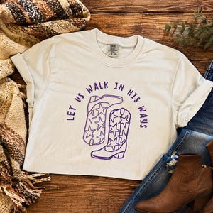 Puede incluir: Una camiseta de manga corta de color claro con un gr&aacute;fico morado de dos botas vaqueras decoradas con patrones de estrellas y el texto "LET US WALK IN HIS WAYS" arqueado sobre las botas. La camiseta tiene cuello redondo y etiqueta.