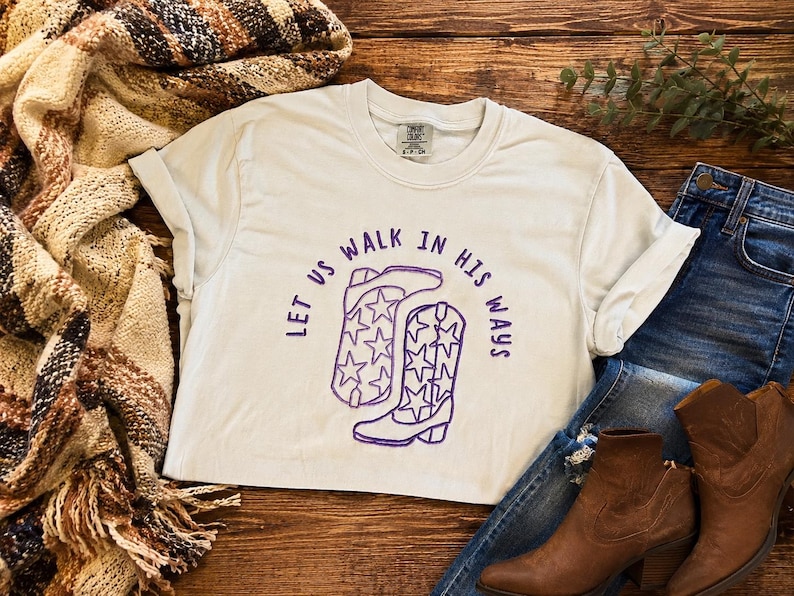 Puede incluir: Una camiseta de manga corta de color claro con un gr&aacute;fico morado de dos botas vaqueras decoradas con patrones de estrellas y el texto "LET US WALK IN HIS WAYS" arqueado sobre las botas. La camiseta tiene cuello redondo y etiqueta.