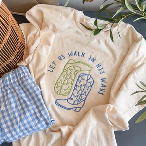 Puede incluir: Una camiseta de color crema con un dise&ntilde;o bordado de dos botas vaqueras, una en verde claro y otra en azul, ambas decoradas con patrones de estrellas. El texto "LET US WALK IN HIS WAYS" est&aacute; bordado en azul alrededor de las botas.