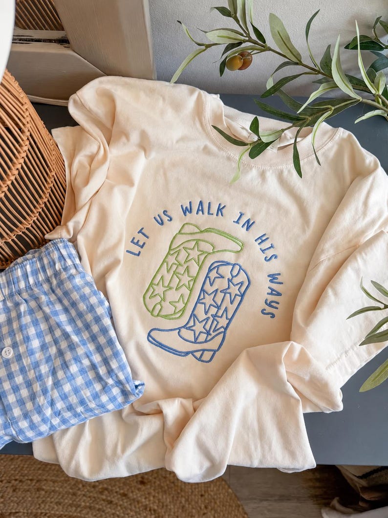 Puede incluir: Una camiseta de color crema con un dise&ntilde;o bordado de dos botas vaqueras, una en verde claro y otra en azul, ambas decoradas con patrones de estrellas. El texto "LET US WALK IN HIS WAYS" est&aacute; bordado en azul alrededor de las botas.