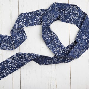 Op de afbeelding: Een marineblauwe bandana sjaal met een wit paisley patroon. De sjaal is gemaakt van een zachte stof en ligt op een wit houten oppervlak. De sjaal is een veelzijdig accessoire.