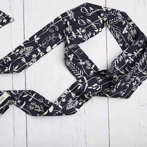 Op de afbeelding: Een marineblauwe sjaal met een wit bloemmotief. De sjaal is gemaakt van een zachte stof en is op een decoratieve manier gevouwen. Het label luidt "BONGOED". Het wordt gepresenteerd op een wit houten oppervlak.