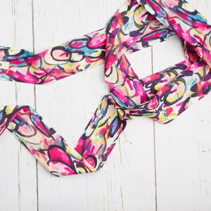 May include: A colorful silk scarf with a vibrant abstract pattern in shades of pink, blue, yellow, and black. The scarf is tied in a knot, and the word "BONGOOD" is visible on a small tag. The scarf is lying on a white wooden surface.