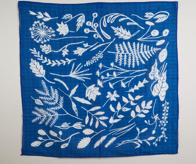 May include: A square, blue bandana with a white botanical design. The design features various leaves, sprigs, and floral elements. The bandana is made of a soft, lightweight fabric.