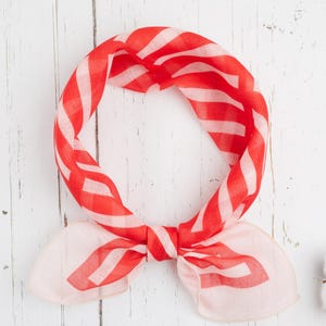 May include: A red and white striped bandana, knotted at the bottom, lies on a white wooden surface. The bandana's pattern is a series of bold, horizontal stripes. A few cotton buds are in the lower right corner.