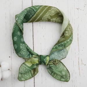Puede incluir: Un pañuelo verde con estampado de cachemira, anudado en forma circular. El pañuelo presenta tonos verdes y blancos, con un diseño floral detallado. El fondo es una superficie de madera blanca, con un tallo de algodón en la esquina inferior izquierda.