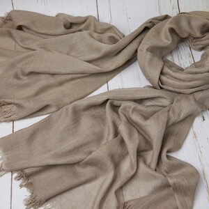May include: A taupe-colored scarf with fringed edges, draped on a white wooden surface. The scarf is made of a soft, flowing fabric. Yellow flowers are in the upper right corner. The scarf is a versatile accessory.