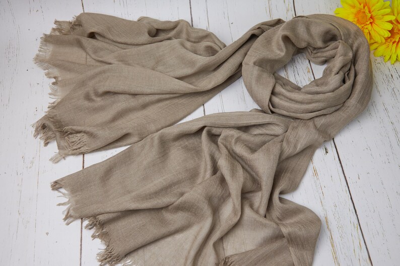 May include: A taupe-colored scarf with fringed edges, draped on a white wooden surface. The scarf is made of a soft, flowing fabric. Yellow flowers are in the upper right corner. The scarf is a versatile accessory.