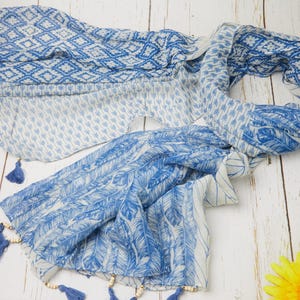 May include: A blue and white patterned scarf with tassel accents. The scarf features various designs, including diamond and feather patterns. The tassels are blue and the scarf is draped on a white wooden surface.