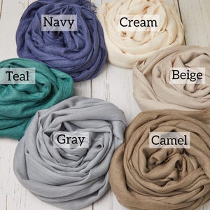 May include: Six rolled scarves in various colors: navy, cream, teal, beige, gray, and camel. Each scarf is labeled with its color name. The scarves are arranged on a white wooden surface.