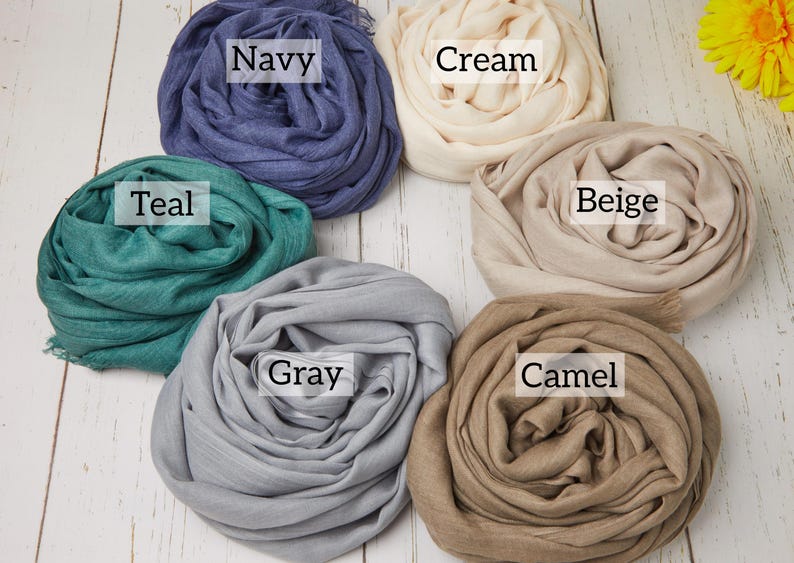 May include: Six rolled scarves in various colors: navy, cream, teal, beige, gray, and camel. Each scarf is labeled with its color name. The scarves are arranged on a white wooden surface.