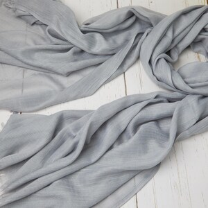 May include: A light gray scarf with fringed edges, draped on a white wooden surface. The scarf is made of a soft, flowing fabric, with visible texture and folds. A yellow flower is visible in the background.