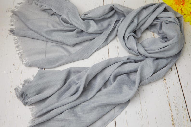 May include: A light gray scarf with fringed edges, draped on a white wooden surface. The scarf is made of a soft, flowing fabric, with visible texture and folds. A yellow flower is visible in the background.