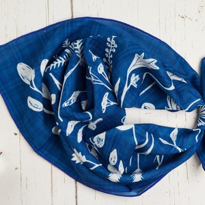 May include: A square, indigo blue scarf with a white floral pattern. The scarf is knotted and lies on a white wooden surface. The floral design includes various leaves and flower shapes. A cotton branch is in the lower left corner.